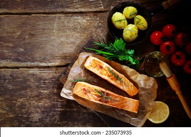 Overhead View Of Delicious Grilled Savory Salmon Cutlets With Ingredients Including A Bouquet Garni Of Fresh Herbs, Olive Oil, Tomatoes, Baby Potatoes And Lemon On A Rustic Wood Table With Copyspace