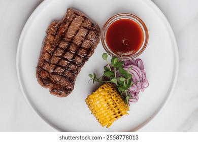 Overhead view of a delicious grilled striploin steak with grilled corn on the cob, pickled red onions, and a small bowl of barbecue sauce, placed on tabletop. Restaurant or cafe menu, cookbook recipe - Powered by Shutterstock