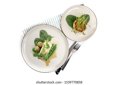 Overhead View Of A Dinner Setting With  A Baked Filet Of Halibut Fish And A Fried Crab Cake Garnished With Spinach And Pea Shoots Isolated On White