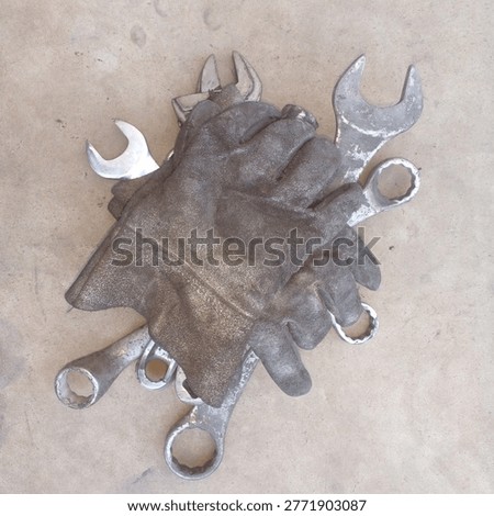 Overhead view of dirty, worn work gloves gently resting on a pile of various metal wrenches on a concrete floor. Authentic and powerful concept for craftsmanship, love for tools, mastery of trade, complete set, readiness for work, the mechanic's touch, dignity of skilled labor, and the bond between a worker and his instruments.