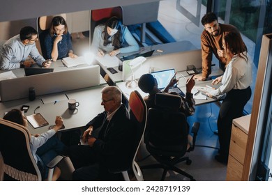 Overhead view of a diverse business team working late, collaborating around a table in a modern office setting. - Powered by Shutterstock