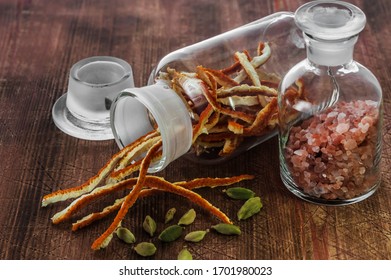 An Overhead View Of Dried Strips Of Orange Zest Into A Glass Vial With A Ground Lid, Green Cardamom Grains And Pink Salt Crystals Into A Glass Vial