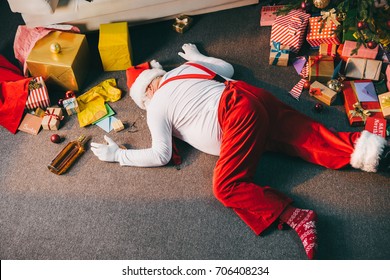 Overhead View Of Drunk Bad Santa Lying On Floor With Bottle Of Whiskey And Wrapped Christmas Gifts