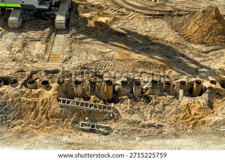 Overhead view of dynamic construction site, revealing heavy excavator tracks, freshly excavated earth, and series of large steel piling casings installed for new foundations.