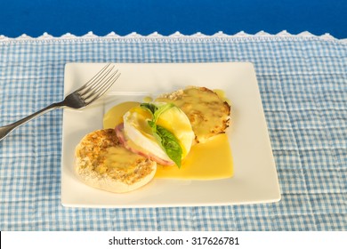 Overhead View Of Eggs Benedict On Square White Plate On Blue Gingham Place Mat.  Garnished With Basil Leaf