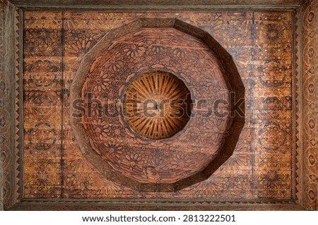 An overhead view of the exceptional painted cedar ceiling of the Mosque Qaddour al-Alami shows a large central circular medallion with radiating geometric arabesques, surrounded by dense painted panels in red, orange, and gold.