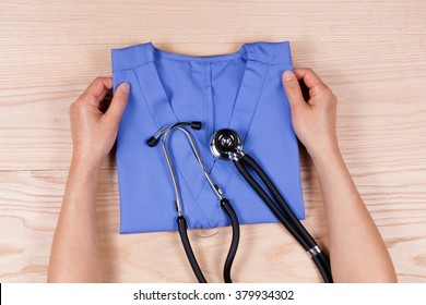 Overhead View Of Female Hands Folding Medical Scrub Shirt With Stethoscope On Wooden Desktop. 

