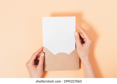Overhead view female hands holding open brown paper envelope with blank white sheet template, beige background