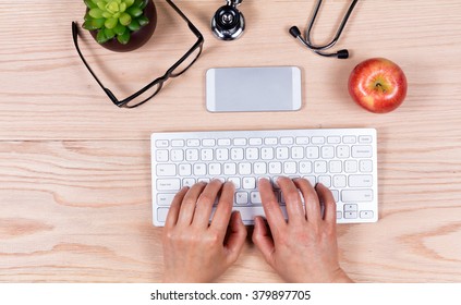 Overhead View Of Female Hands Typing On Computer Keyboard With Medical Items On Wooden Desktop. 