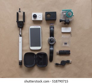  Overhead View. Flat Lay, Top View  Of Essential Vacation Item. Travel Concept Background On Craft Paper With Selfie Stick, Watch, Action Camera, Phone, Glasses, Case, Xiaomi