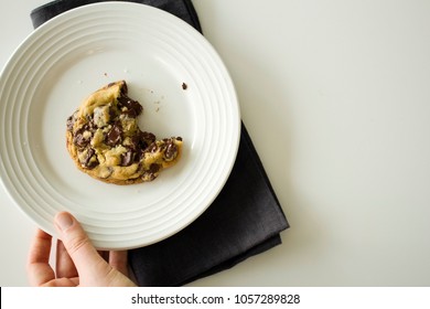 Overhead View Of Fresh Chocolate Chip Cookie With Bite Mark Hand Holding Plate And Dark Linen