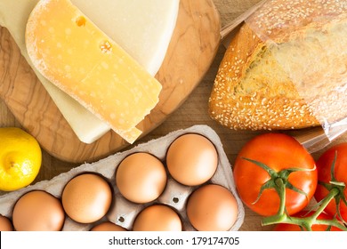 Overhead View Of Fresh Groceries In The Kitchen With A Cardboard Carton Of Free Range Eggs, Grape Tomatoes On The Vine, Cheese, Crusty Seed Bread And A Lemon