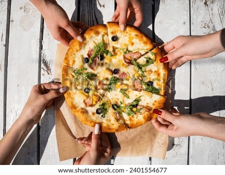 Overhead view of a freshly baked artisan pizza with arugula, olives, corn, and ham, with people's hands taking slices, indicating a communal meal
