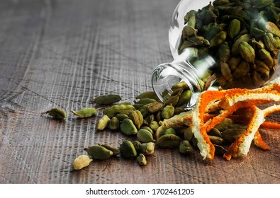 An Overhead View Of Green Cardamom Grains Running Out Of A Glass Bottle And Dried Strips Of Orange Zest On A Wooden Table