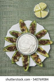 An Overhead View Of A Group Of Meat And Cheese Stuffed Jalapeno Pepper Halves On A Plate Surrounding A Bowl Of Sour Cream For Dipping And A Bowl Of Quartered Lime Wedges Off To The Side. 