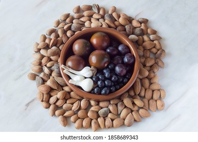 Overhead View Of A Handmade Clay Bowl With Organic Products, Garlic Heads, Tomatoes, Blueberries, And Black Grapes Surrounded By Almonds With Shells On A Rustic Marble Table. Healthy Food.