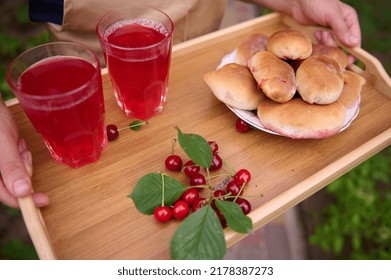 Overhead View Of The Hands Of A Housewife Holding A Serving Tray With Freshly Baked Cherry Pies And Drinking Glasses With Refreshing Compote From Cherries, Standing In The Backyard Of A Private House