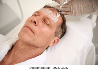 Overhead View Of Hands In Medical Gloves Of Aesthetician Holding A Make-up Brush And Applying Beauty Cosmetic Moisturizing Cleansing Product On Mature Man's Face. Male Beauty Procedures Concept At Spa