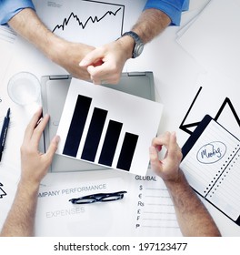 An Overhead View Of The Hands Of Two Men Studying A Chart Over A Closed Laptop.