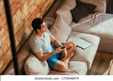 Overhead View Of Handsome Man Holding Remote Controller And Watching Tv On Sofa With Laptop During Quarantine