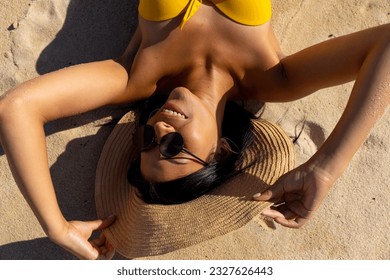 Overhead view of happy biracial woman in sunhat and sunglasses lying on sand at sunny beach smiling. Sunbathing, summer, free time, relaxation, lifestyle and vacation, unaltered. - Powered by Shutterstock