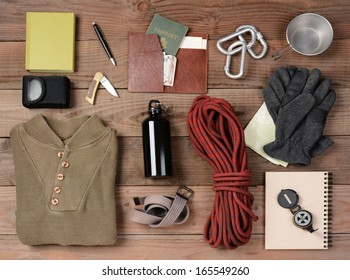 Overhead view of hiking gear laid out for a backpacking trip on a rustic wood floor. Items include, rope, gloves, sweater, carabiners  book, belt, cup, passport, wallet, canteen, compass, money, map,