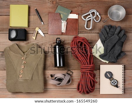 Overhead view of hiking gear laid out for a backpacking trip on a rustic wood floor. Items include, rope, gloves, sweater, carabiners  book, belt, cup, passport, wallet, canteen, compass, money, map,