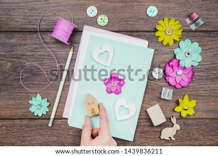 An overhead view of human hand making greeting card with flowers and wooden house block on desk