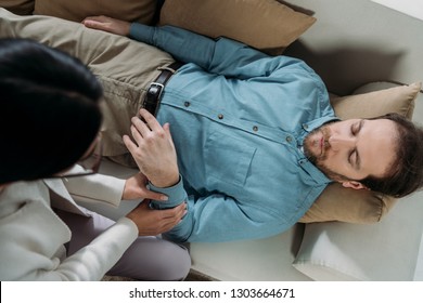 Overhead View Of Hypnosis Holding Wrist Of Bearded Man With Closed Eyes Lying On Couch