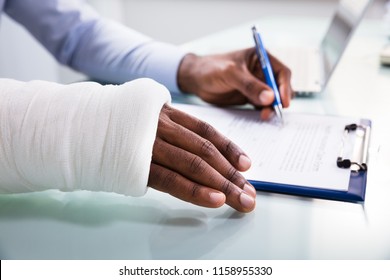 Overhead View Of Injured Man With Bandage Hand Filling Insurance Claim Form On Clipboard