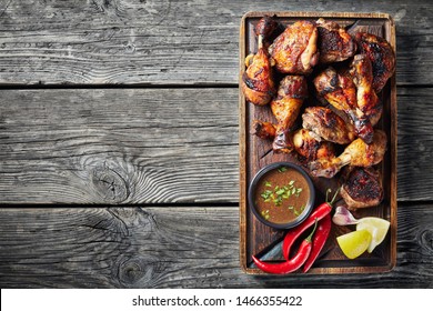 overhead view of Jerk Chicken on a rude board with sauce and lemon on a rustic wooden table, horizontal view from above, close-up, flatlay, copy space