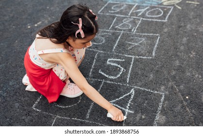 Overhead View Of A Little Girl Drawing With Chalk Hopscotch On Playground. Child Playing The Game Outside. The Kid Draws Hopscotch On The Pavement. Activities For Children.