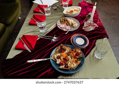 Overhead view of a long dining table covered with a sage-green cloth and a draped red velvet runner, set with red napkins, cutlery, and water glasses. A lit white taper candle in a glass holder sits near shared dishes including a plate of olives, cheese, and cured meats, a basket of bread, and a blue plate holding slices of savory tart topped with roasted cherry tomatoes.