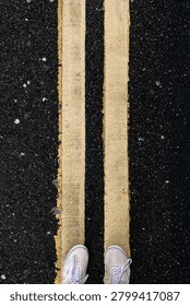 Overhead view looking down at white sneakers standing between double yellow road markings on black asphalt, urban street photography perspective