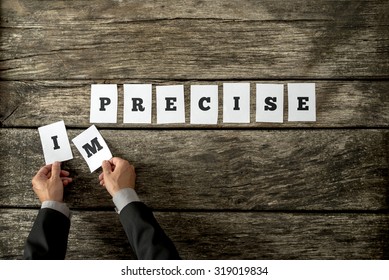 Overhead View Of Male Hands Removing Letters IM From The Word Imprecise Changing It Into Precise Spelled On White Cards Over Aged Wooden Boards.