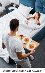 Overhead View Of Man Holding Wooden Tray With Breakfast While Woman Sleeping In Bed