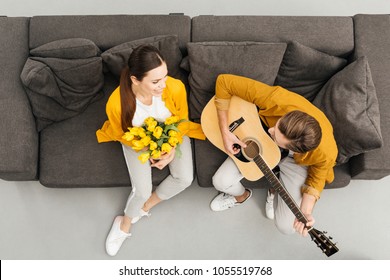 Overhead View Of Man Playing Guitar To His Girlfriend While She Holding Bouquet On Couch At Home