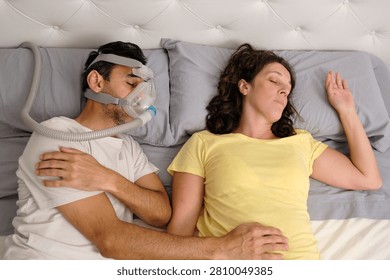 Overhead view of a man using a CPAP machine for sleep apnea while sleeping next to his partner in a comfortable bedroom. Concept of sleep disorder treatment, healthcare, and healthy relationships