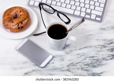 Overhead View Marble Counter Top With Spoon In Dark Coffee, Computer Keyboard, Cell Phone, Reading Glasses, And Bagel On Plate. Work At Home Concept. Selective Focus On Spoon In Cup. 