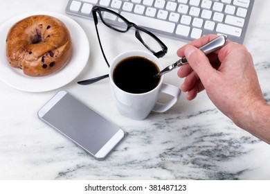 Overhead View Marble Kitchen Counter Top With Male Hand Stirring Coffee With Spoon, Computer Keyboard, Cell Phone, Reading Glasses, And Bagel On Plate. Work At Home Concept. 