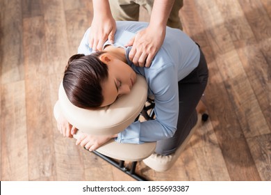 Overhead view of masseur massaging shoulders of brunette businesswoman with closed eyes, sitting on massage chair