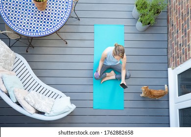 Overhead View Of Mature Woman With Pet Cat At Home Checking Phone After Exercise On Deck