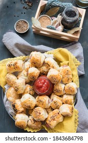 Overhead View Of A Metallic Dish Full Of Baked Sausage Rolls On A Yellow Cloth With A Ketchup Jar In The Middle. A Wooden Basket With Some Spice Jars Inside Is At The Upper Part Of The Image.