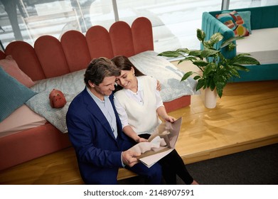 Overhead View Of A Middle Aged Couple In Love Sitting Close To Each Other And Looking Through A Catalog With Upholstered Furniture While Choosing Home Furniture In Shopping Mall. Home Interior Design