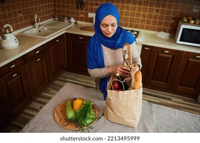 Overhead View Of A Muslim Middle-Eastern Pretty Woman In Blue Hijab, Removing Groceries From A Shopping Bag, In The Kitchen At Home. Healthy Grocery Purchasing. Food Delivery.