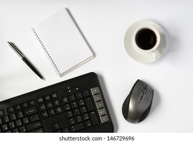 Overhead View Of An Office Work Station With A Wireless Computer Mouse, Keyboard, A Cup Of Espresso Coffee And A Blank Notebook And Pen On A White Surface