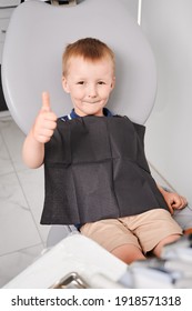 Overhead View On A Little Patient Sitting In Dentist's Chair. Smiling Boy, Wearing Black Bib And Showing Thumb Up At Dentist Office. Vertical Snapshot. Healthcare Concept