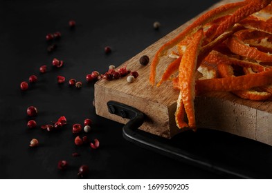 An Overhead View On Strips Of Orange Zest And Red Peppercorns Lying On A Beech Cutting Board With A Black Forged Metal Handle In A Black Background