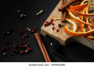 An Overhead View On Strips Of Orange Zest, Cinnamon Sticks And Red Peppercorns Lying On A Beech Cutting Board With A Black Forged Metal Handle In A Black Background