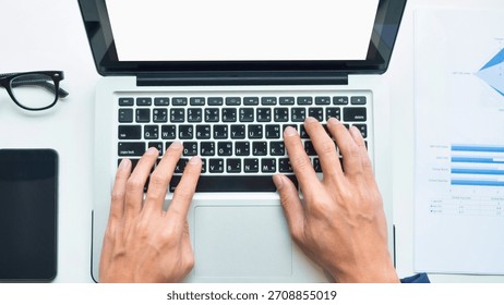 Overhead view of a person's hands typing on a laptop. A tablet, glasses, and charts are also visible.